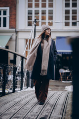 Obraz premium Stylish young woman wearing a warm coat, scarf, and knitted hat walking in a European city street during autumn or winter. Urban lifestyle portrait with cozy fashion, natural light.