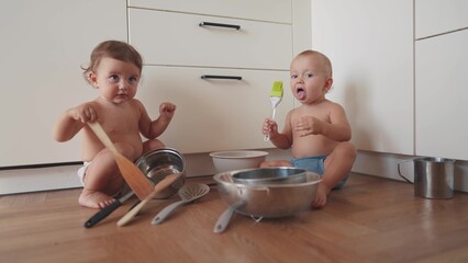 Fototapeta premium baby group playing pots in the kitchen on the floor. happy family kid dream concept. two baby twins scattered pots play dabble sitting on the floor. lifestyle baby scattered dishes pots mess