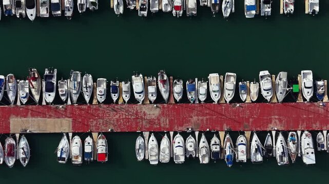 Many boats are parked side by side at a red dock. The scene shows clear water around the boats. This marina is busy with various sizes of boats moored.