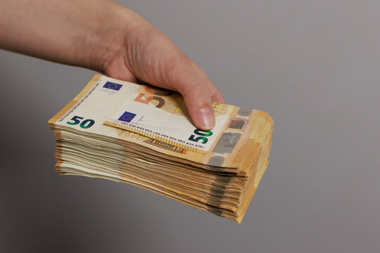 A hand holds a thick stack of 50 euro banknotes against a light, neutral background. The banknotes are neatly stacked, and the distinctive euro design is clearly visible. 