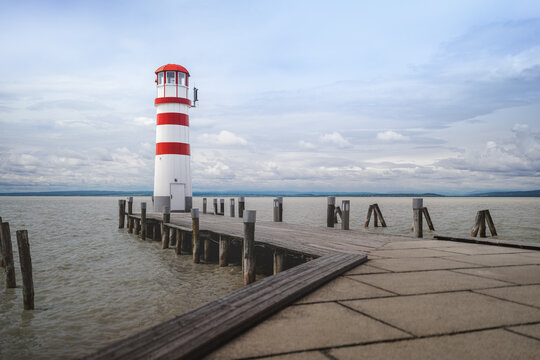 Scenic view of the red and white lighthouse at the end of a wooden pier in Podersdorf am See on Lake Neusiedl, Austria, under a cloudy sky.