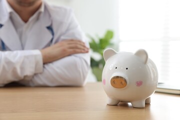 Doctor sitting at wooden table indoors, focus on piggy bank