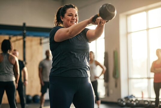 A joyful, body-positive woman smiles while performing a kettlebell swing exercise during a fitness class in a sunlit gym.
