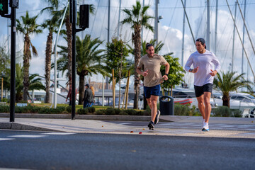 Obraz premium Two men jogging together along a sunny marina promenade with palm trees and boats, enjoying an active urban lifestyle focused on fitness, friendship and outdoor wellbeing