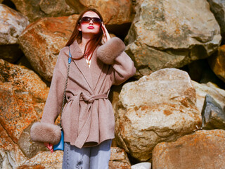 A woman wearing a fur coat and sunglasses stands in front of a pile of rocks. She is enjoying the outdoors and the scenery, beauty and fashion.
