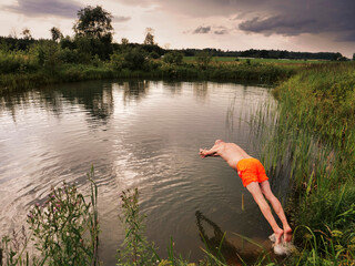 A man is jumping into a pond wearing an orange swimsuit at dusk. The sky is cloudy and the water is calm, colors are muted and calm. Sport and relaxation after a long working day concept.