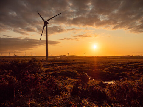 A wind farm with a large wind turbine in the foreground. The sky is orange and the sun is setting. Eco way to generate electricity. Spectacular view. West of Ireland.