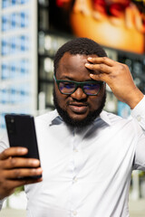 Obraz premium Black male adult receiving upsetting news on his smartphone and feeling worried, frustrated dissatisfied guy in the downtown area. Exploring city center and being unhappy with his phone.