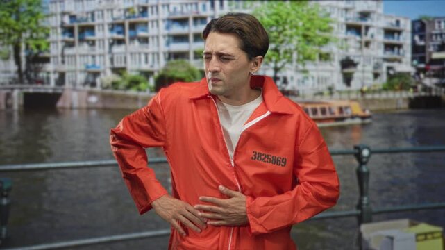 Man prisoner in orange jumpsuit clutching stomach with hands at a building canal railing, inmate number printed on chest; distress.