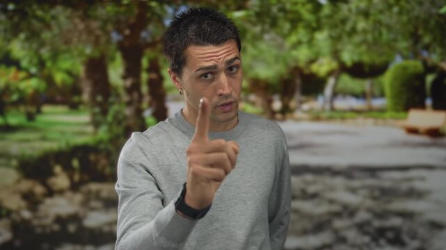 Young hispanic man in grey sweater gesturing seriously with finger pointing in sunny park with green trees and blurred background outdoors during daytime
