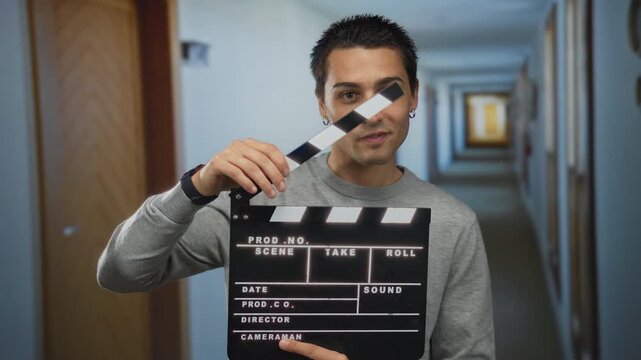 Young hispanic man holding clapperboard in hotel corridor creating film scene enthusiastically indoors representing filmmaking process and cinematic storytelling essence