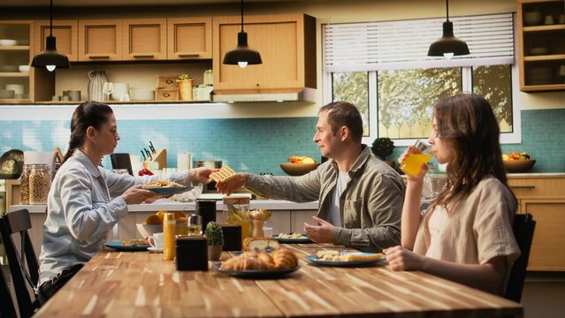 Parents and kids sitting together at breakfast table being served by the mother, serving scrambled eggs with bacon and goods. Cozy scene captures bonding with joy for morning routine.