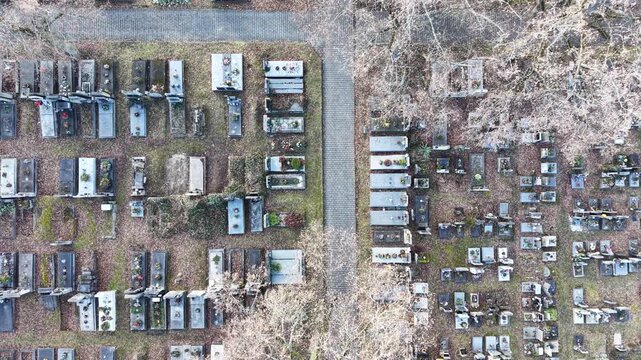 Aerial view of a cemetery. Rows of gravestones are visible. A path runs through the graveyard. Trees are bare. Autumn foliage is present. Peaceful and somber atmosphere