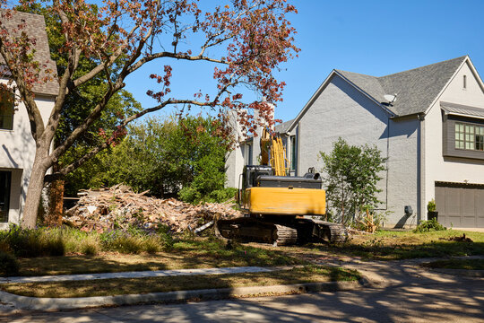 Yellow excavator clearing debris pile from residential lot between modern white brick homes