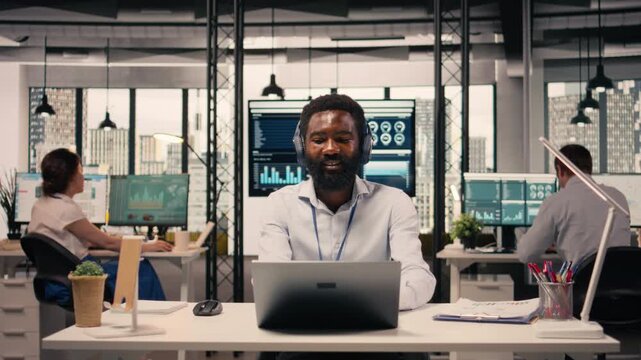African american man in good mood listens to music on headset, working with laptop and combining stress relief with focused business analysis. Multitasking with audio and tasks in office.