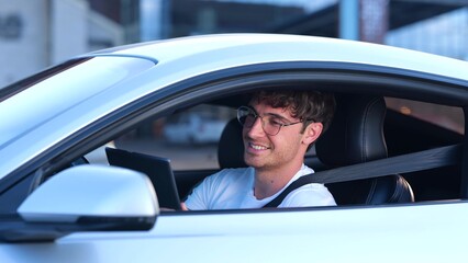 Smiling young man in glasses sitting in the car, using seat belt while working and holding digital tablet. Transport, job, technology and lifestyle concept. Slow motion