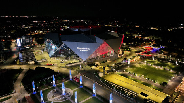 Aerial drone photo of Mercedes-Benz Stadium at night, showcasing the venue illuminated by bright, colorful stadium lights. The striking geometric roof design and modern architecture 