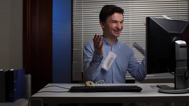 Young man at his computer desk cheering as banknotes rain down, celebrating unexpected financial windfall from online earnings, investment gains, salary bonus and business success