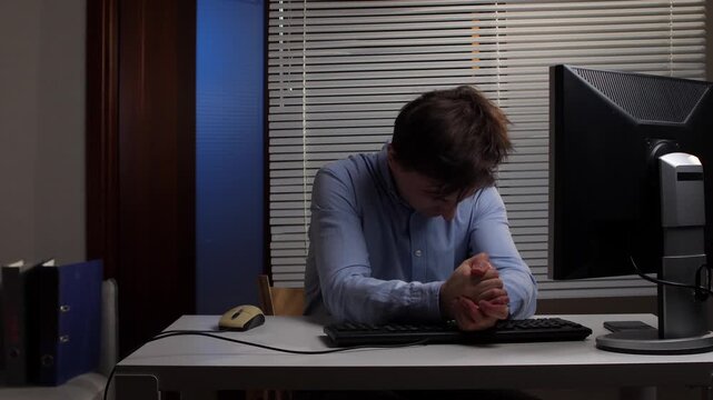 Young man experiencing burnout and pressure, clutching his head in frustration while working overtime on a computer, indicating exhaustion and mental strain in a dimly lit office setting