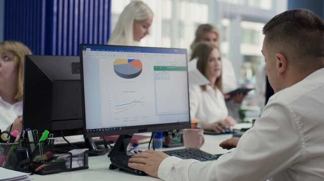 Close-up of an office, shot from behind an employee working at a computer. The monitor shows business charts, with colleagues blurred in the background. 4K.