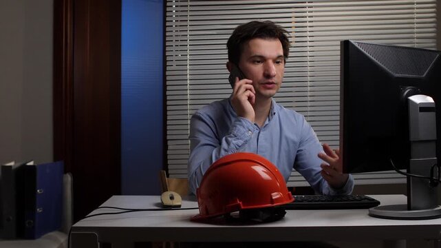 Male construction engineer having a business phone call while sitting at his desk, working on a computer with a safety helmet resting beside him in a modern office