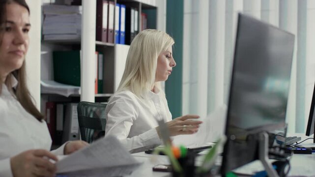 Medium shot, side view, of two office workers working on documents. A blonde woman focuses on the papers in front of her, next to a colleague. 4K.