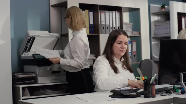 Video sequence: Two Caucasian women performing administrative tasks. One scans or copies documents while the other works at the desk, illustrating efficiency and workflow in a corporate space. 4K.