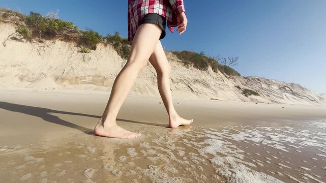 Close up of a woman walking barefoot on sandy beach near rocks and dunes. Steps in wet sand. Summer vacation, relaxation, freedom, seaside lifestyle, and nature journey
