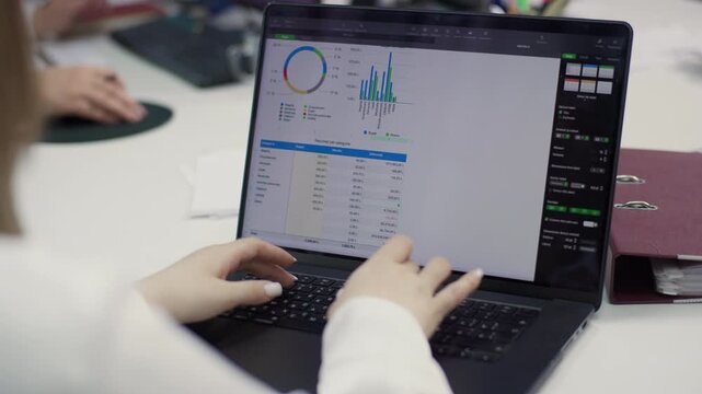 A close-up of a desk, showing the hands of an employee taking notes on a document containing graphs and data. A computer keyboard and monitor are visible. 4K.