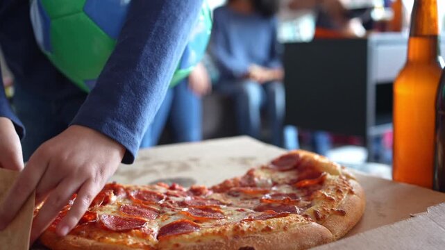 Close up of greasy pizza slices on the table as a little girl reaches in for a piece during soccer game night. Junk food spread while family and friends watch the match