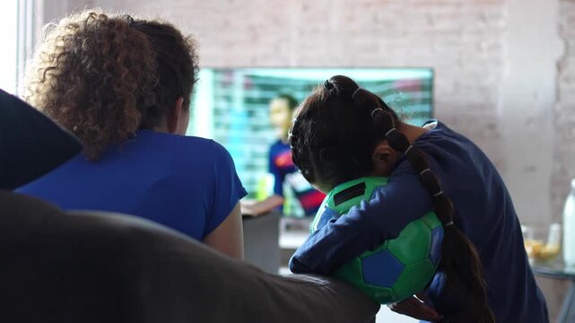 Sad girl hugging a soccer ball while a goal is celebrated on TV behind her. Football and soccer disappointment at home, sports loss, family and friends watching match