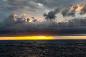 Sunrise over Mediterranean Sea with vibrant clouds in Sardinia, Italy. Golden light reflects on calm waters. Coastline stretches with soft morning light.