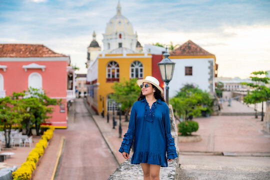 Young smiling Latina woman enjoys a peaceful morning on the colonial walls of Cartagena, Colombia. Wears a stylish blue dress, sneakers, straw hat. The backdrop glows with vibrant architecture