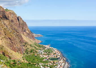 Madeira cliffside village. Steep rock face towers above terraced homes and green plots. Bright blue...