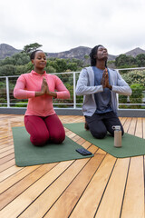 African American couple in sportswear kneeling, meditating on deck with mats, phone, bottle nearby
