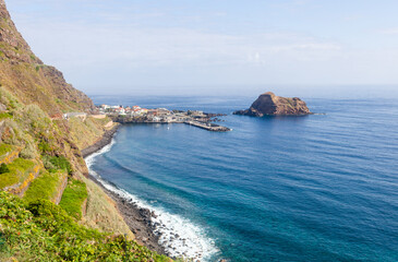 Obraz premium Porto Moniz coastline. Rocky promontory and small harbour sit beside deep blue Atlantic waters. Volcanic islet rises offshore near lava rock shore.