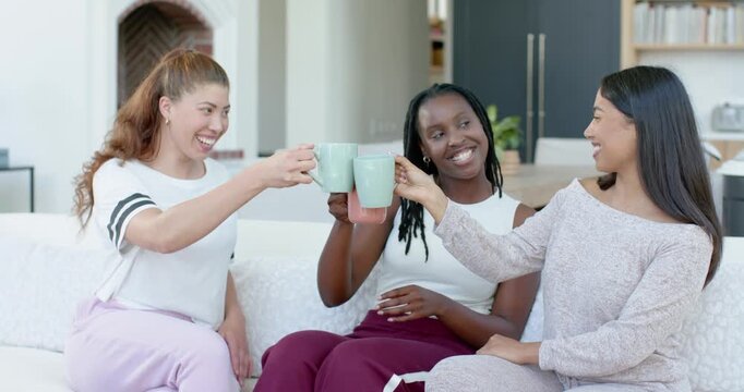 Diverse female friends raising and clinking mugs, laughing, cradling pink mug catching up on sofa