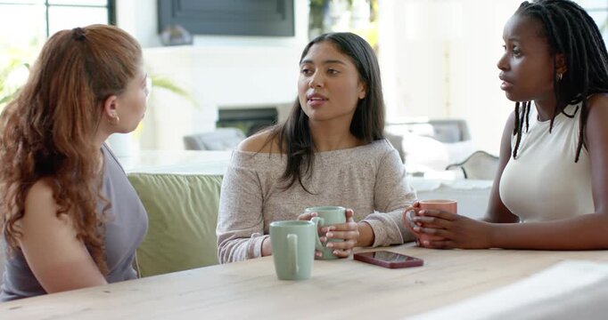 Three women friends sitting at home left friend speaking center woman holding green mug expressing