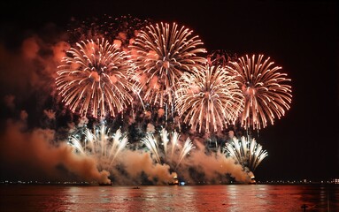 Fireworks explode over water at night, casting vibrant colors on the dark sky and reflective surface.