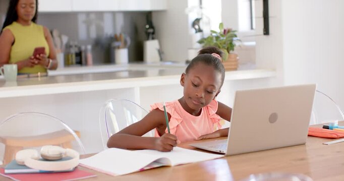 African family youth girl doing homework using laptop in kitchen while phone pinging mom checking