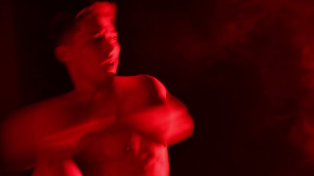 Athletic man in boxing gloves training in red light and smoke against black background. Camera moving forward