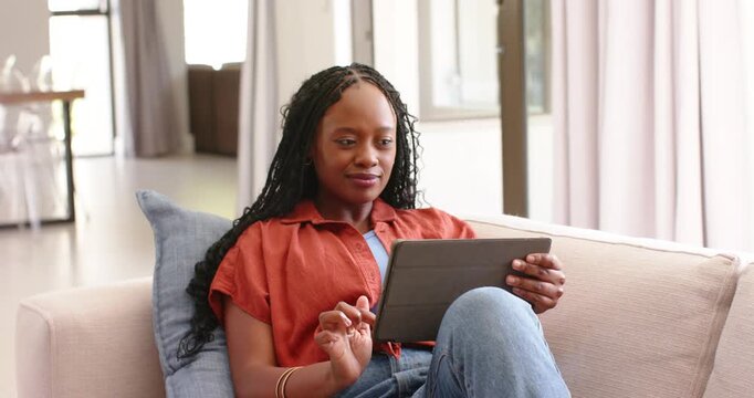 Adult woman sitting on sofa with blue pillow at home holding tablet, tapping and swiping screen