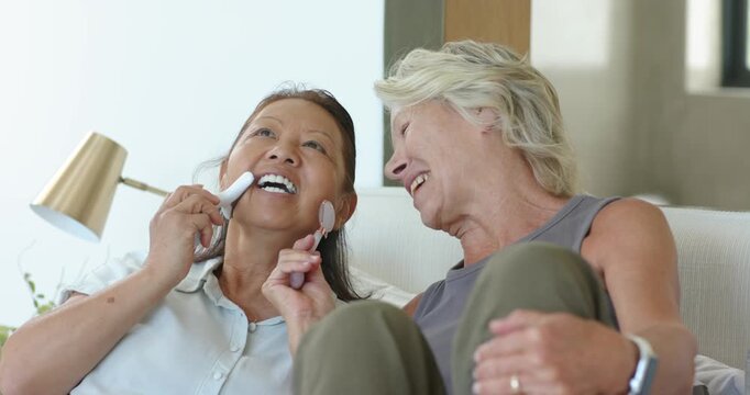 Senior Asian and African American women smiling using rollers and LED tool on sofa sharing skincare