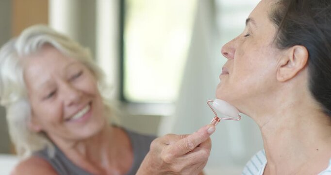 2 female friends on sofa, recipient tilting to expose jaw, friend rolling rose quartz for skincare