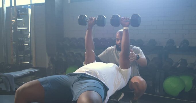 African male trainer spotting trainee at gym bench with dumbbells lowering, pressing for power