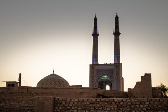 Jameh Mosque of Yazd, Iran, at dusk, with twin blue tiled minarets, patterned dome and monumental iwan portal above the old city walls, icon of Islamic and Persian architecture. in desert city center.