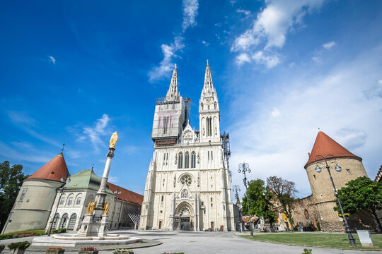 Zagrebacka katedrala, Zagreb Cathedral, seen from Kaptol in the city center with twin spires and restoration scaffolding, framed by historic buildings and a Marian column under a blue sky in Croatia.