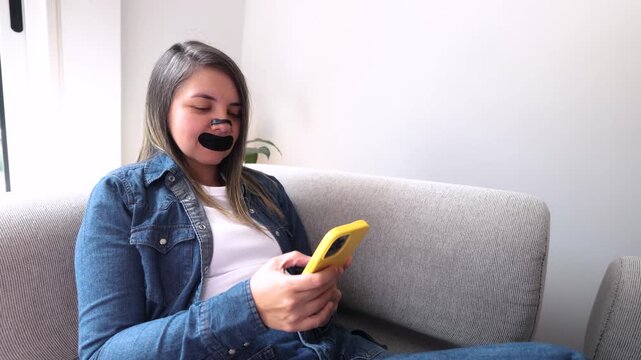 Latina woman using mobile phone on sofa at home with nasal strip and mouth tape &ndash; Biohacking mindfulness and wellness lifestyle