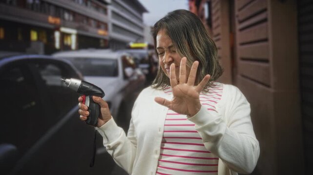 Woman holding cordless screwdriver with palm out stop gesture and squinting face on busy city street; caution safety.