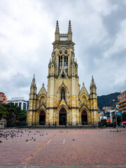 Naklejka premium Front view of the historic Nuestra Senora de Lourdes Basilica with stone facade and tall spires under a cloudy sky at a public square with pigeons in Bogota, Colombia.